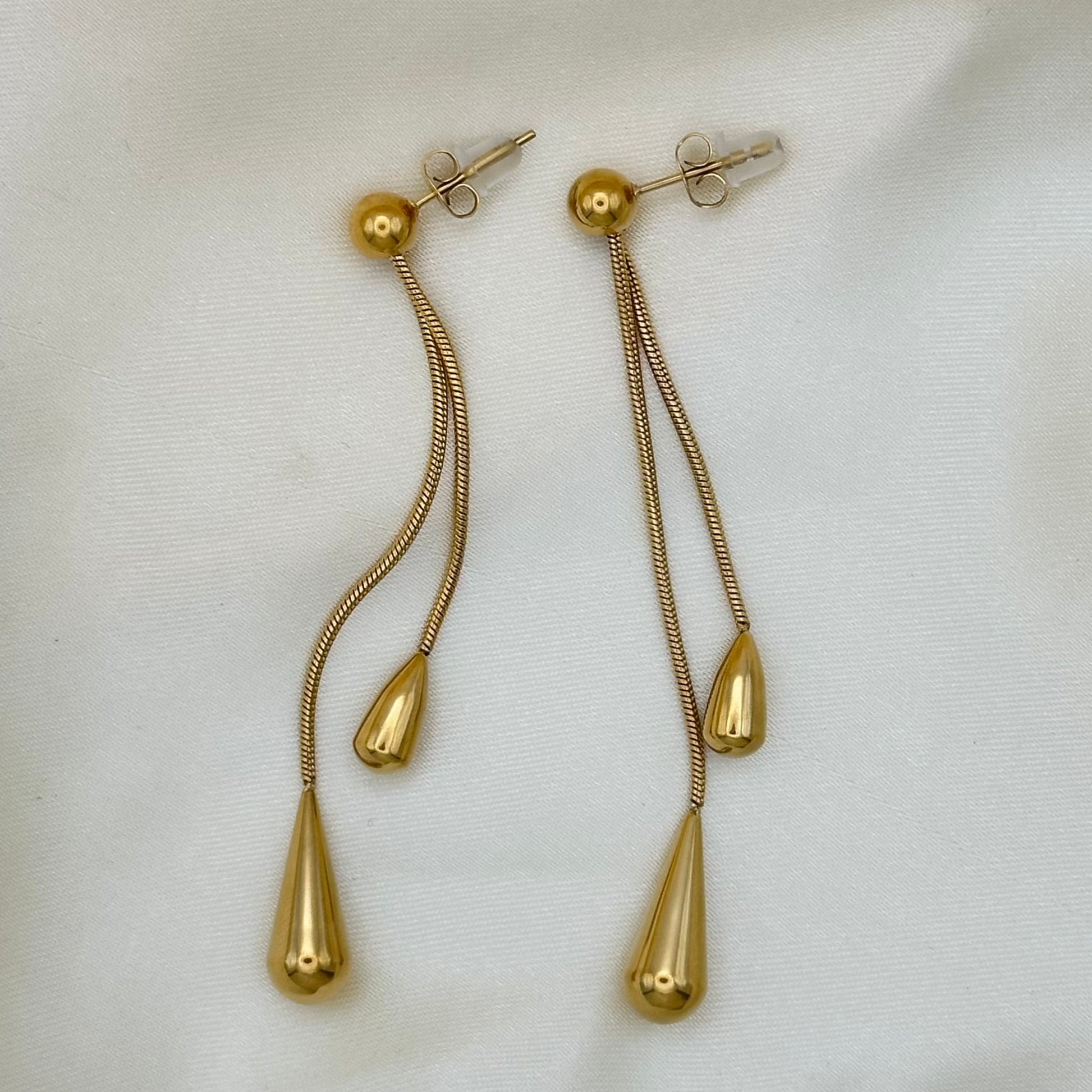 A pair of gold dangle earrings with textured snake-chain drops, teardrop-shaped beads, and a larger teardrop pendant, displayed on a white textured fabric background.