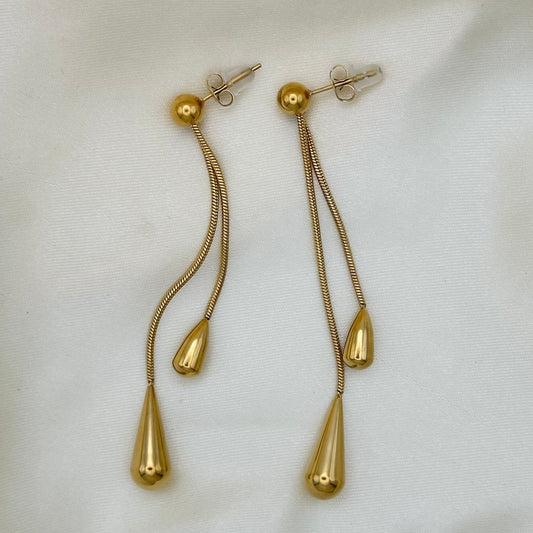 A pair of gold dangle earrings with textured snake-chain drops, teardrop-shaped beads, and a larger teardrop pendant, displayed on a white textured fabric background.