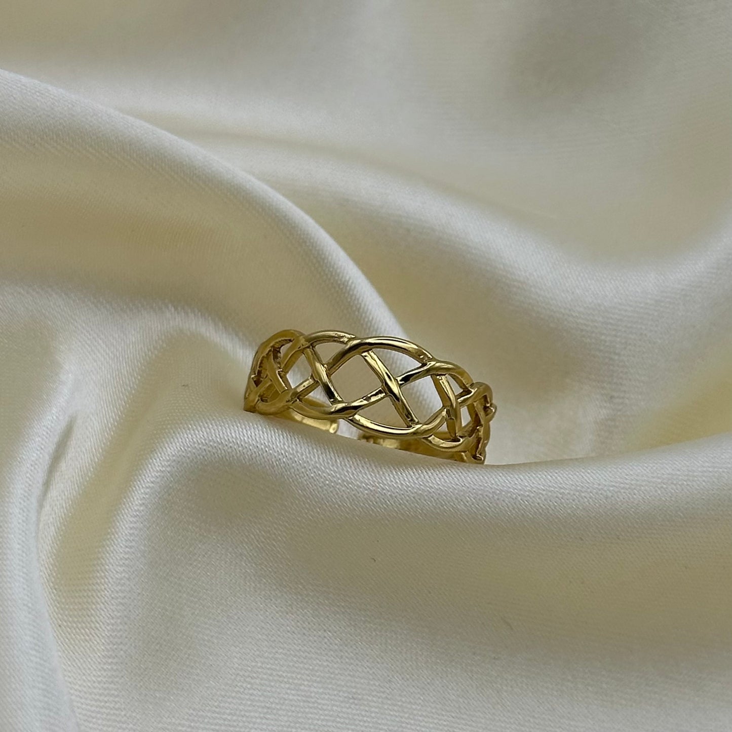 A gold ring with an openwork interlaced knot design, resting on draped cream-colored satin fabric.