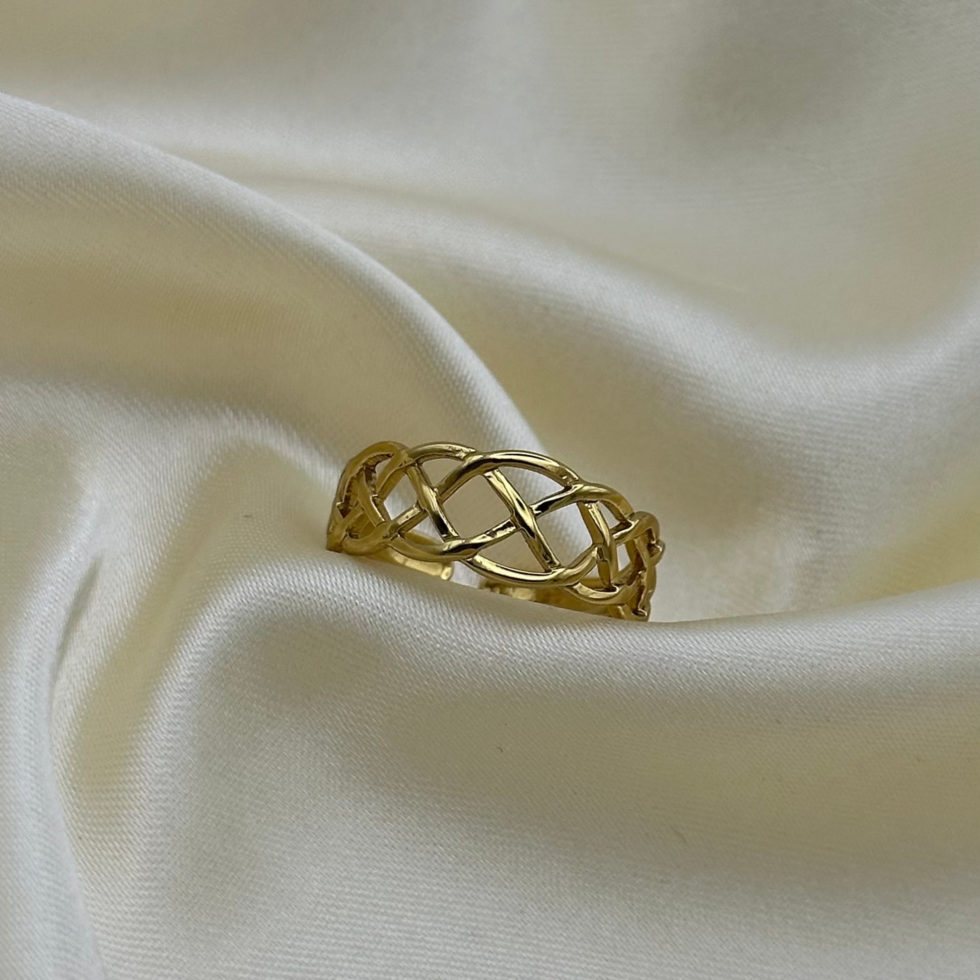 A gold ring with an openwork interlaced knot design, resting on draped cream-colored satin fabric.