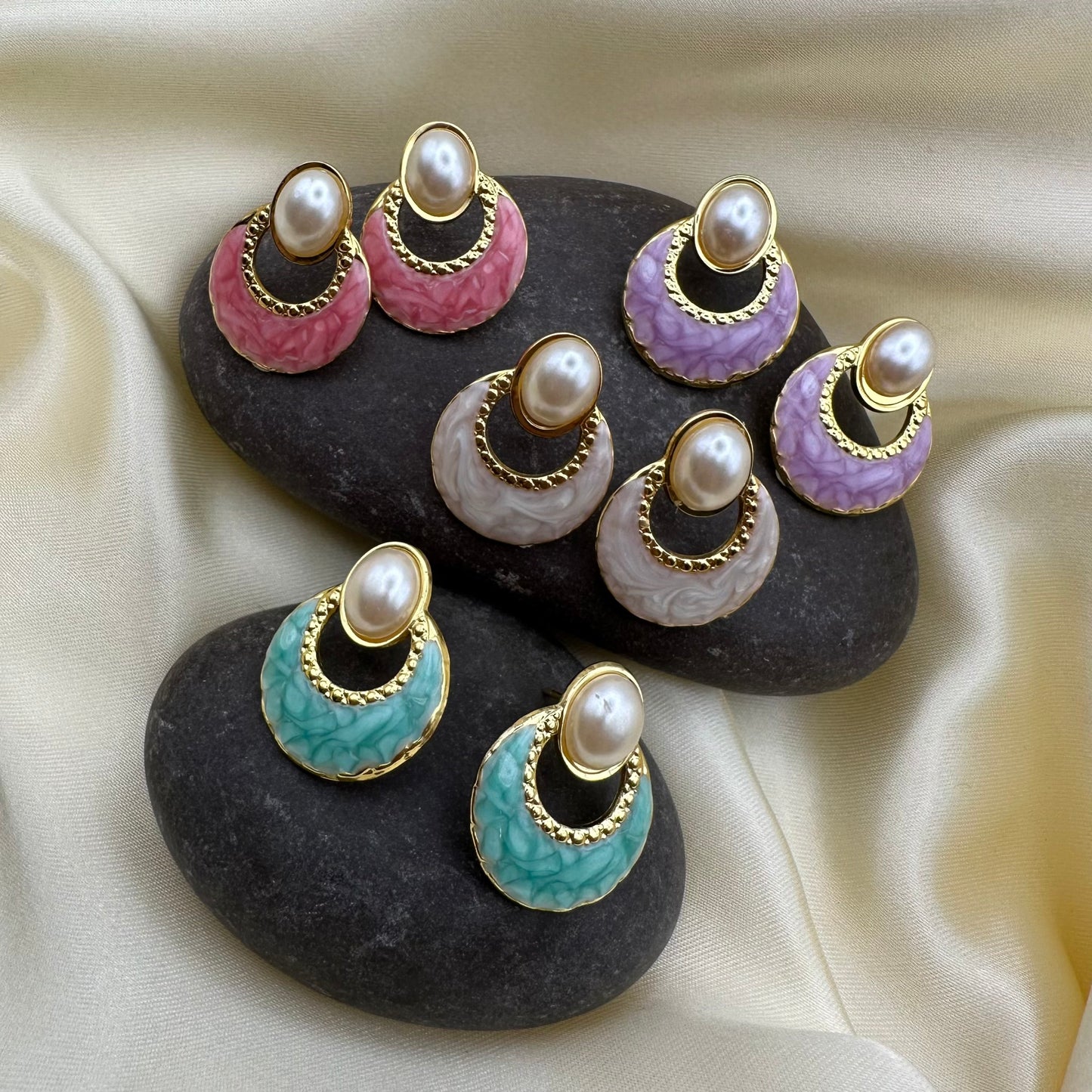 Four pairs of drop earrings displayed on dark stones over cream satin fabric. Each earring has a round pearl-like top, a gold-tone rim, and a curved enamel or stone inlay in pink, lavender, pale gray, mint green, or turquoise.