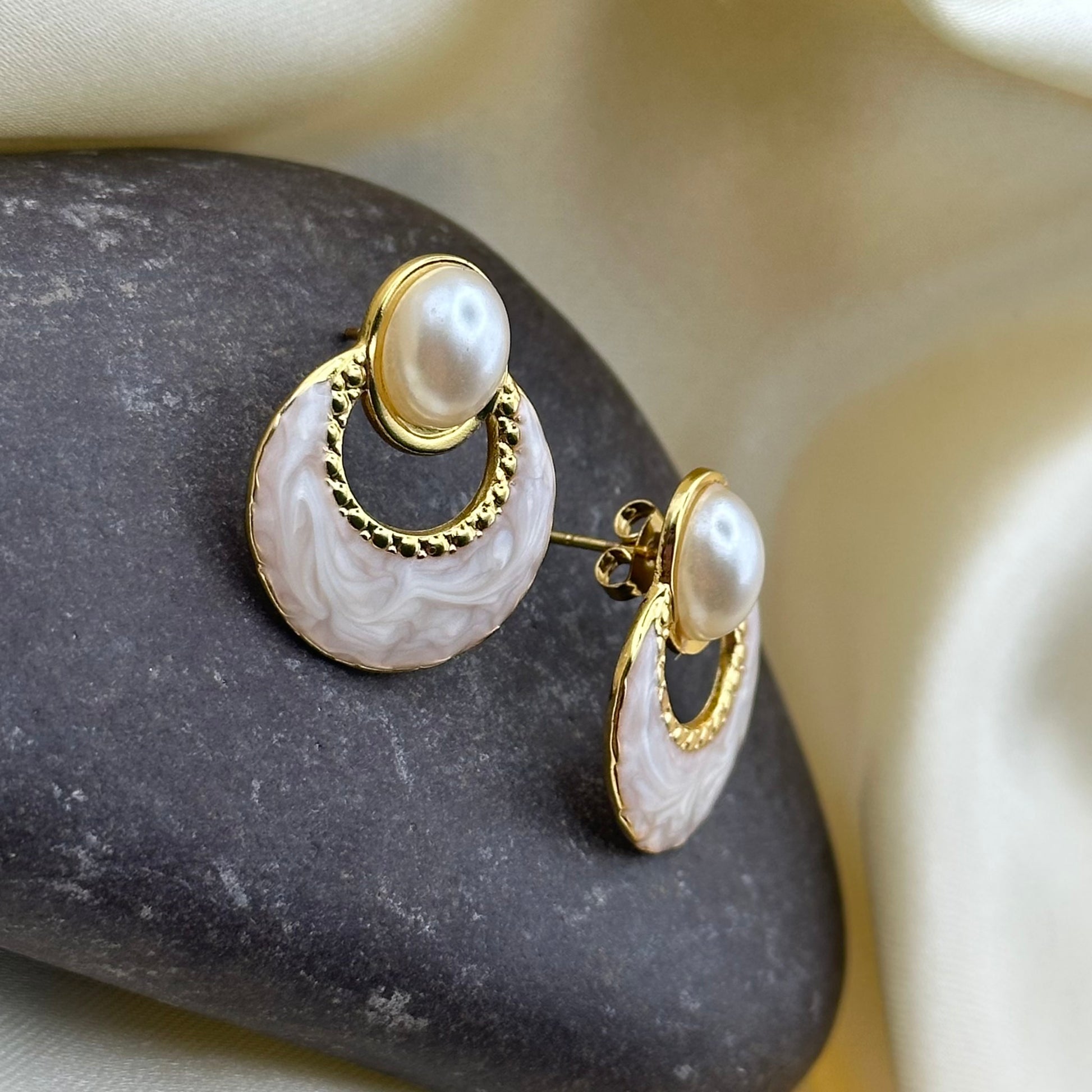 A pair of gold and pearl drop earrings with a circular mother-of-pearl disc, displayed on a dark stone against a cream-colored fabric background.