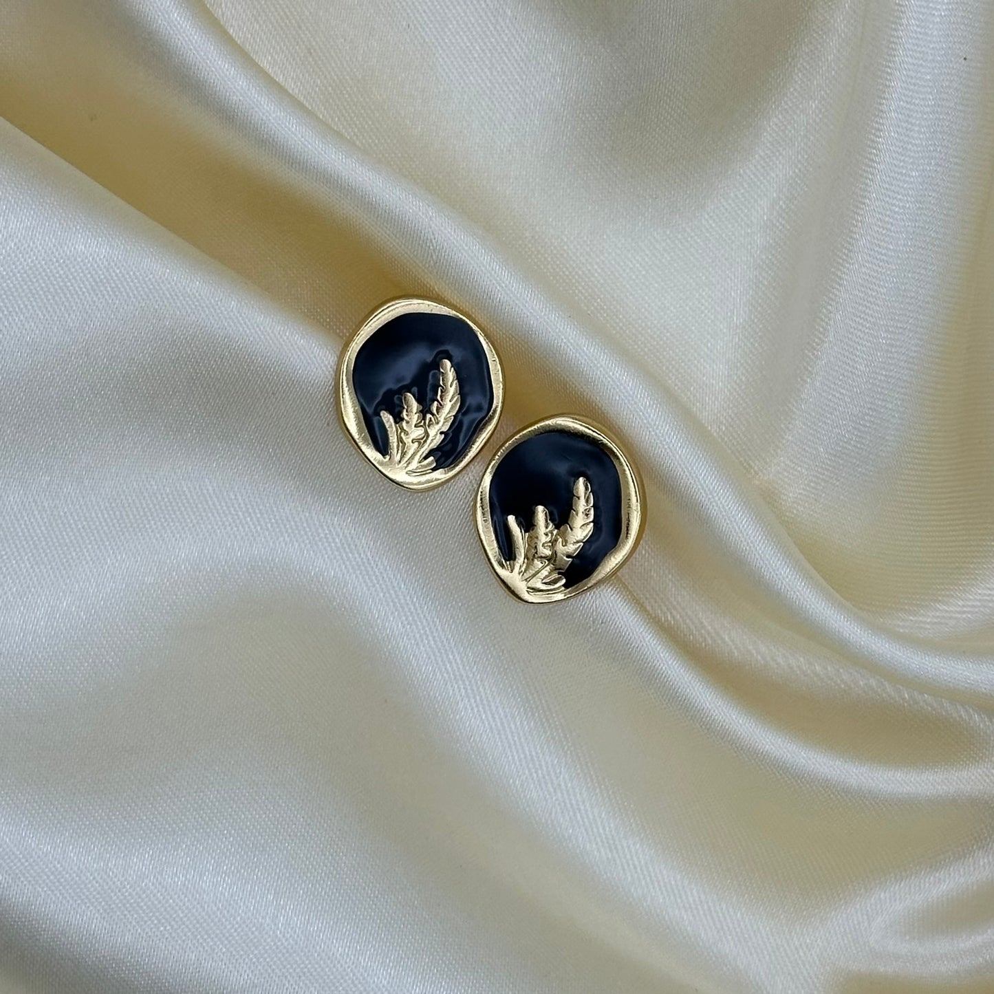 A pair of gold-toned oval stud earrings with a black enamel center and a raised wheat stalk motif, displayed on draped cream satin fabric.