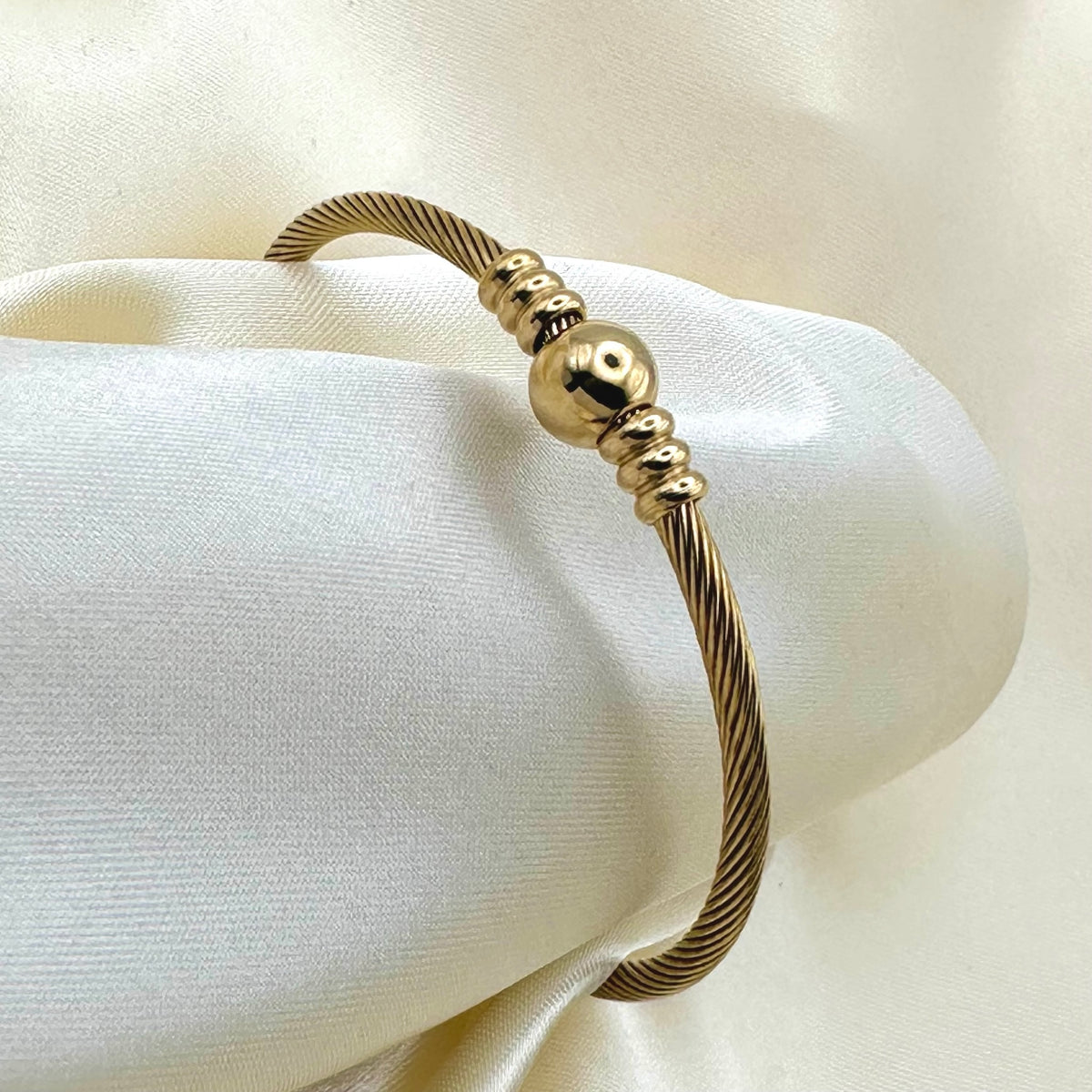 A gold cable-wire cuff bracelet with a central polished bead and fluted bead accents, displayed on draped cream satin fabric.