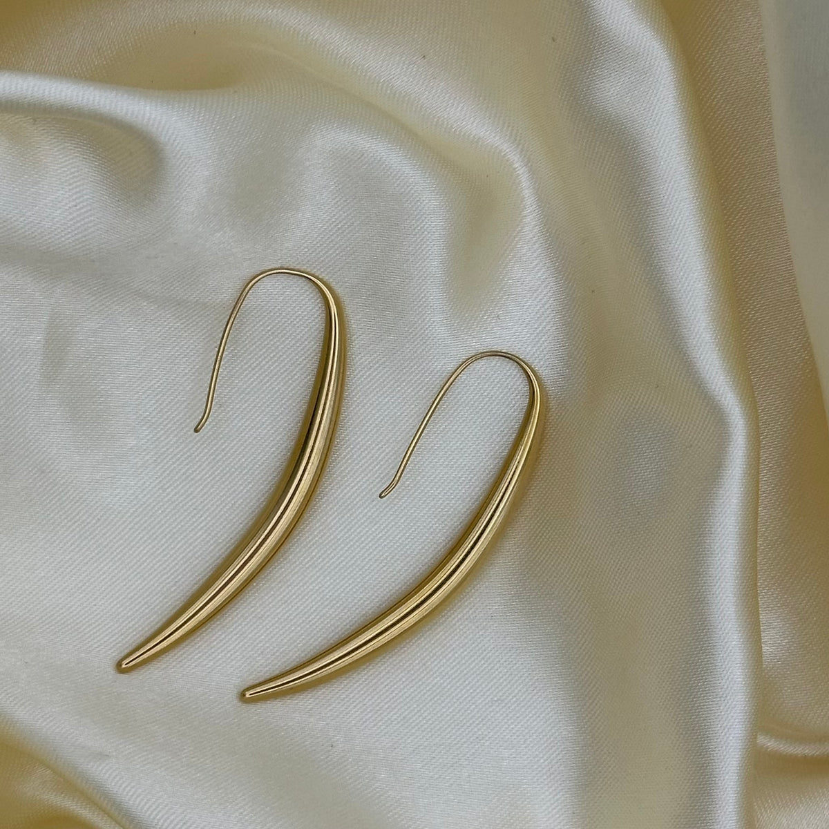 A pair of gold-toned, curved drop earrings with a smooth finish, displayed on a draped cream satin fabric background.