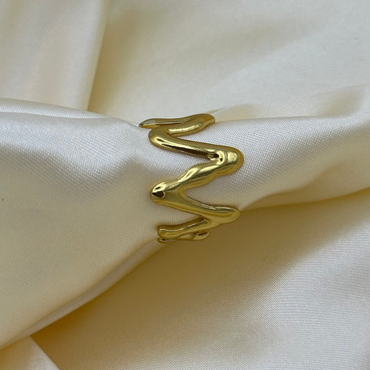 A gold-colored, abstract zigzag open ring displayed on a cream-colored satin fabric.