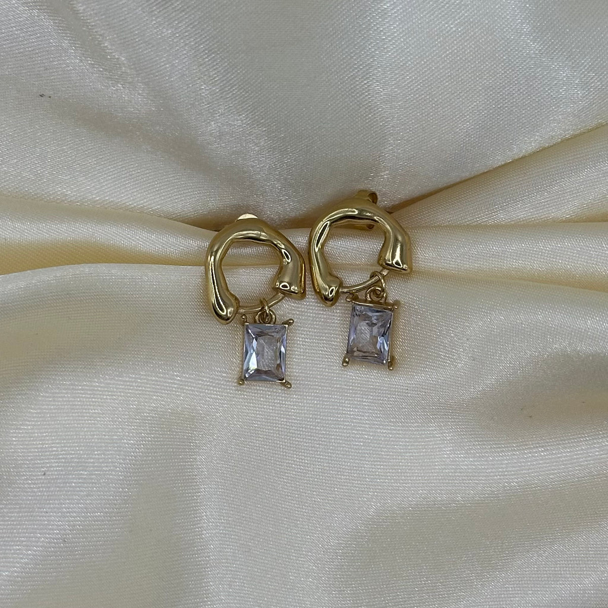 A pair of gold huggie hoop earrings with rectangular clear gemstone drops, displayed on a cream-colored satin fabric background.