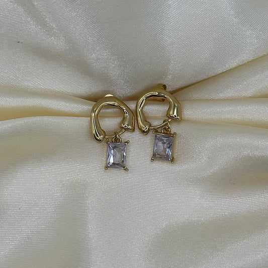 A pair of gold huggie hoop earrings with rectangular clear gemstone drops, displayed on a cream-colored satin fabric background.