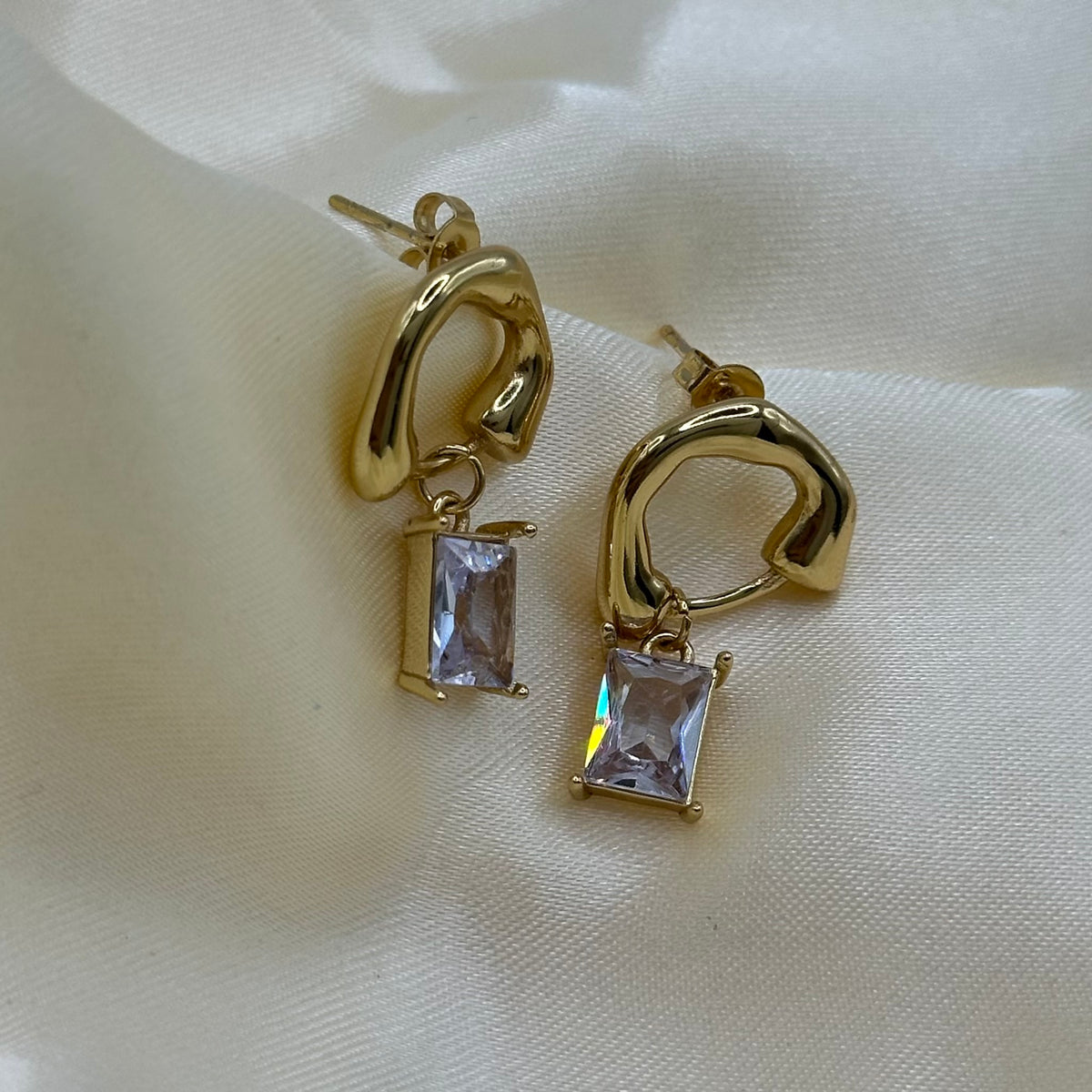 A pair of gold huggie hoop earrings with rectangular clear crystal drops, displayed on a soft, draped cream-colored fabric background.