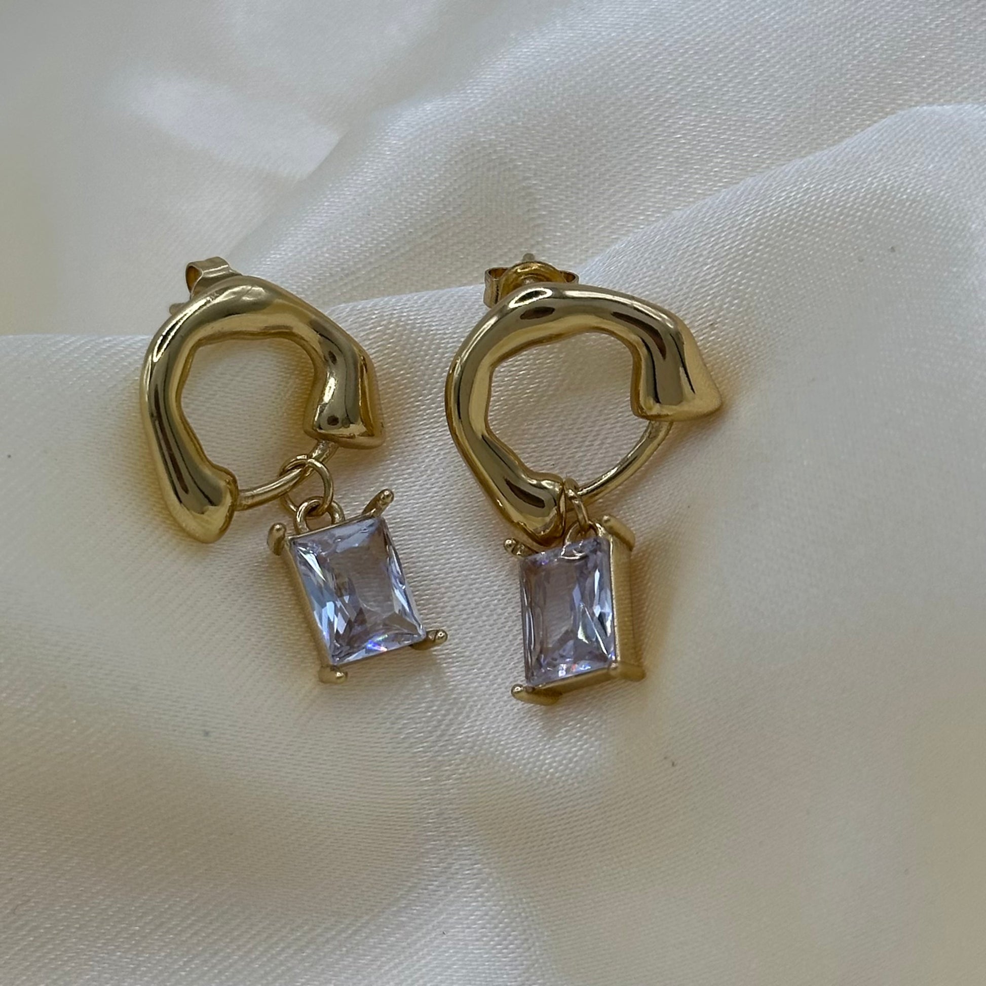 A pair of gold huggie hoop earrings with rectangular clear crystal drops, displayed on a soft cream-colored fabric background.