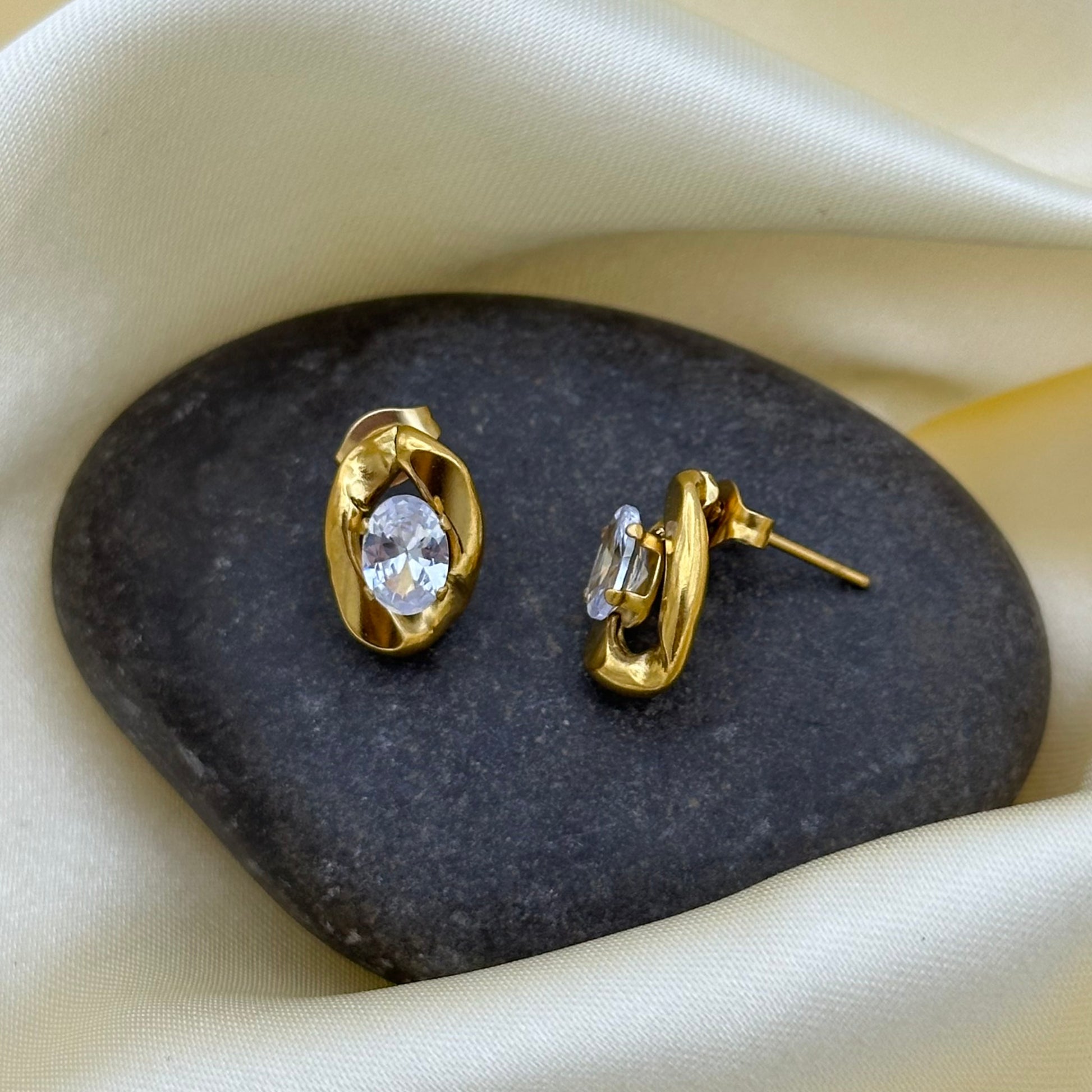 A pair of gold-toned twisted wrap stud earrings with oval clear gemstones, displayed on a dark stone against a draped cream satin fabric background.