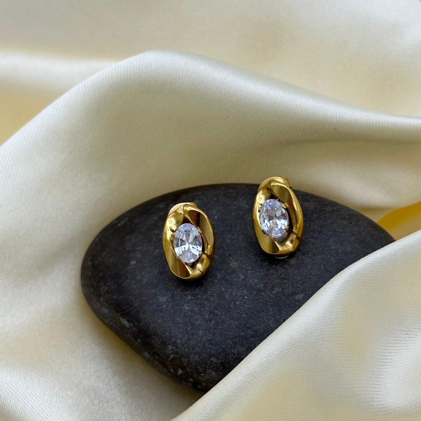 A pair of gold-toned oval stud earrings with clear marquise-cut gemstones, displayed on a dark stone against draped cream satin fabric.