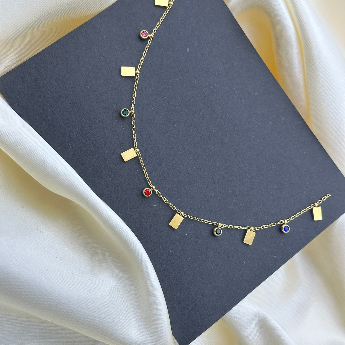 A delicate gold chain necklace with small gold bar and circle charms, some adorned with red, green, blue, and black stones, displayed on a black card over cream-colored draped fabric.