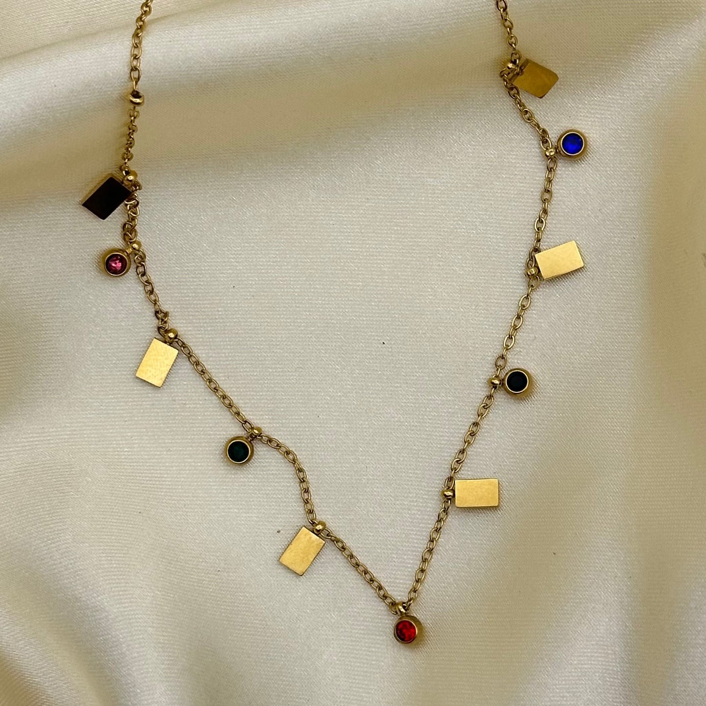 A gold chain necklace with geometric charms, including rectangular and circular pieces, some set with small colored stones in red, blue, and black, displayed on a cream satin fabric background.