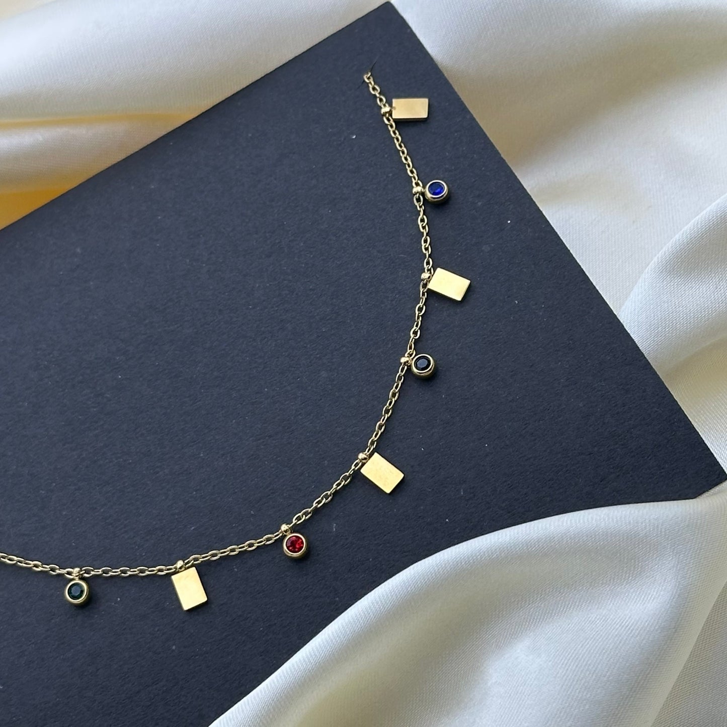 A delicate gold chain necklace with small rectangular gold charms and five round bezel-set gemstone pendants (green, red, black, blue and pink) displayed on a dark charcoal card over white satin fabric.
