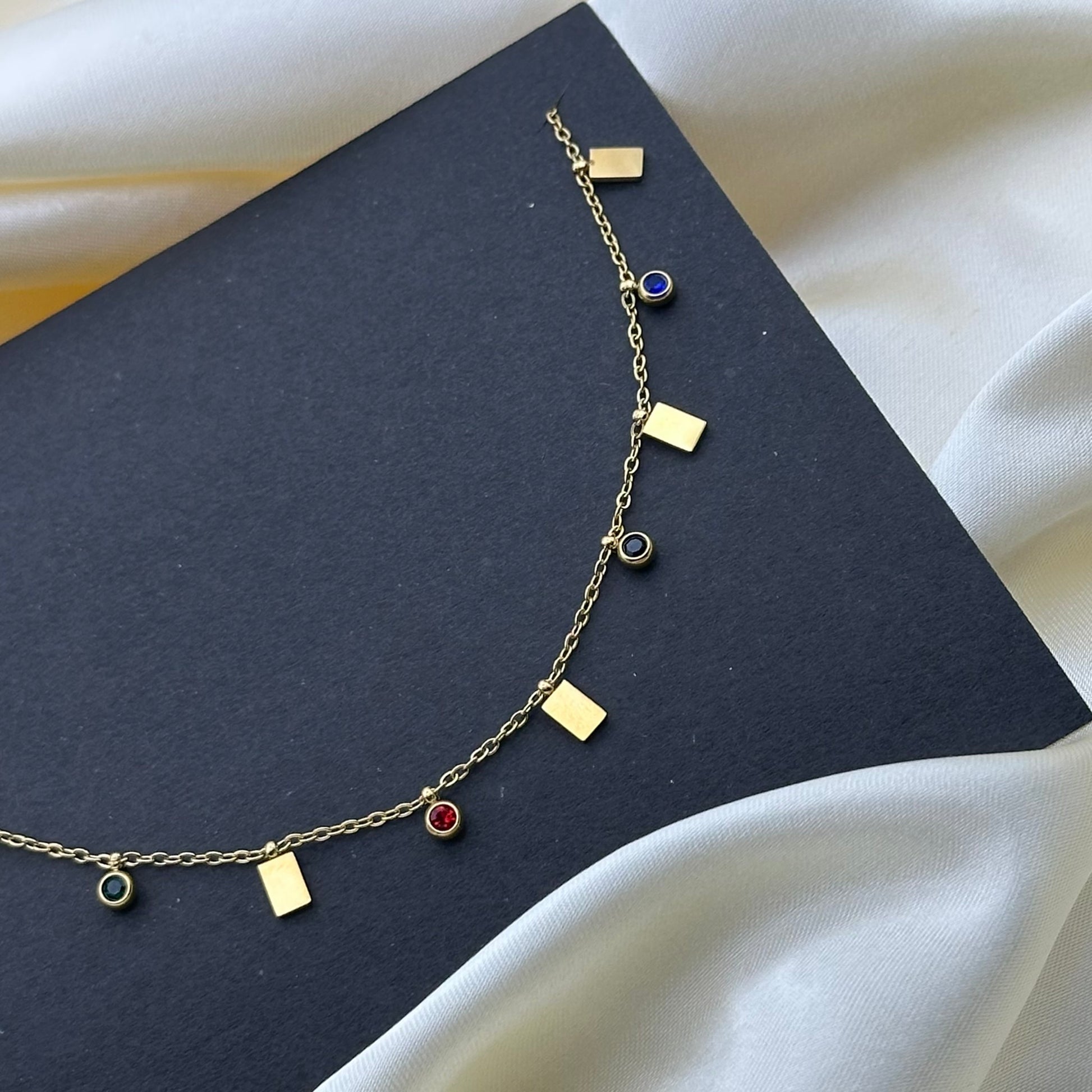 A delicate gold chain necklace with small rectangular gold charms and five round bezel-set gemstone pendants (green, red, black, blue and pink) displayed on a dark charcoal card over white satin fabric.