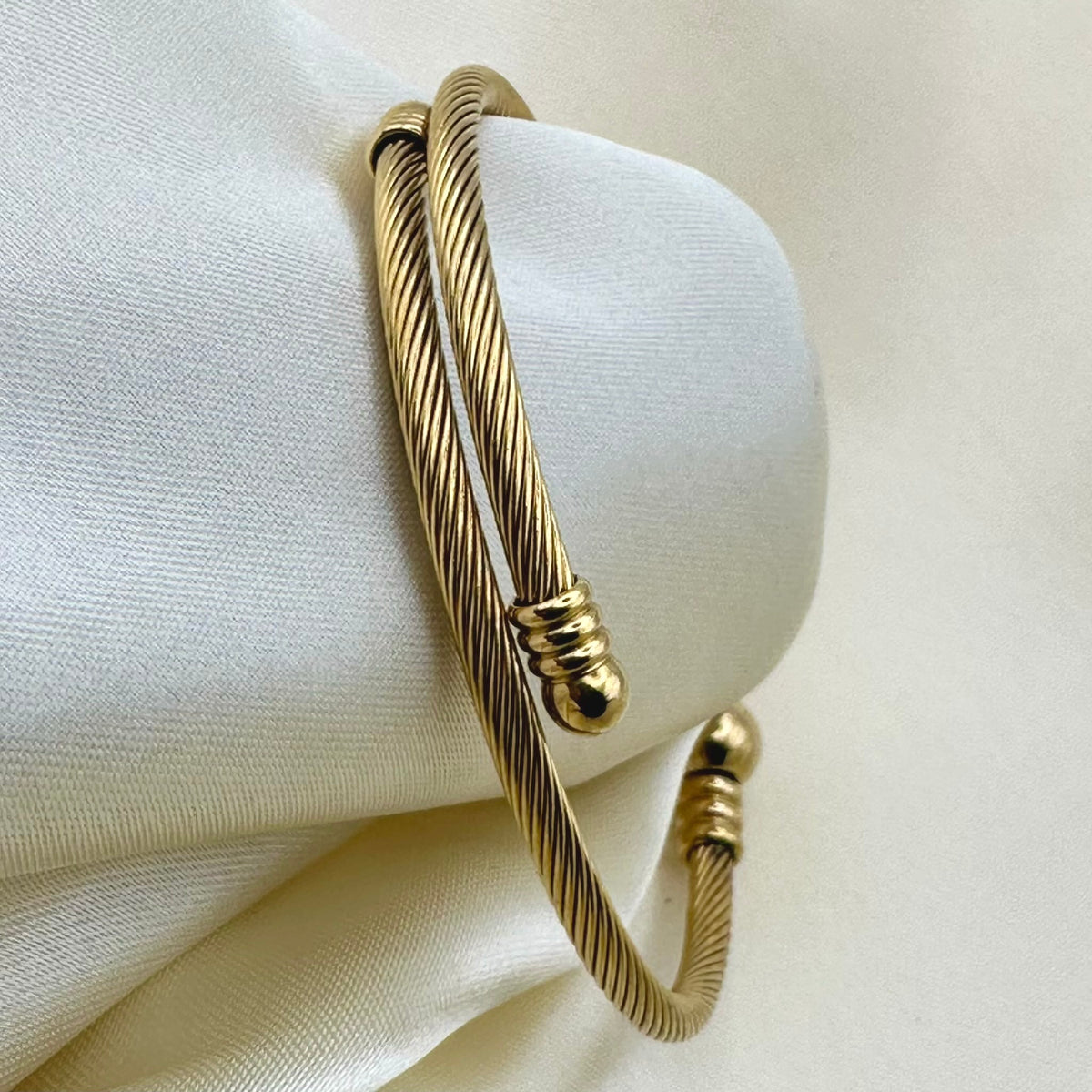 A gold-toned twisted cable bangle bracelet displayed on a soft cream fabric surface.