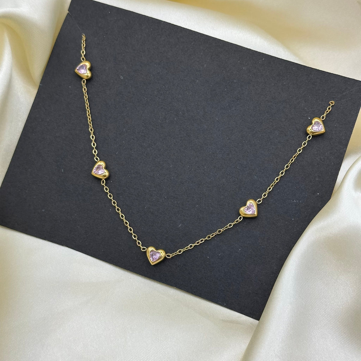A delicate gold chain necklace with small heart-shaped charms, each featuring a light pink inlay, displayed on a black card over cream satin fabric.