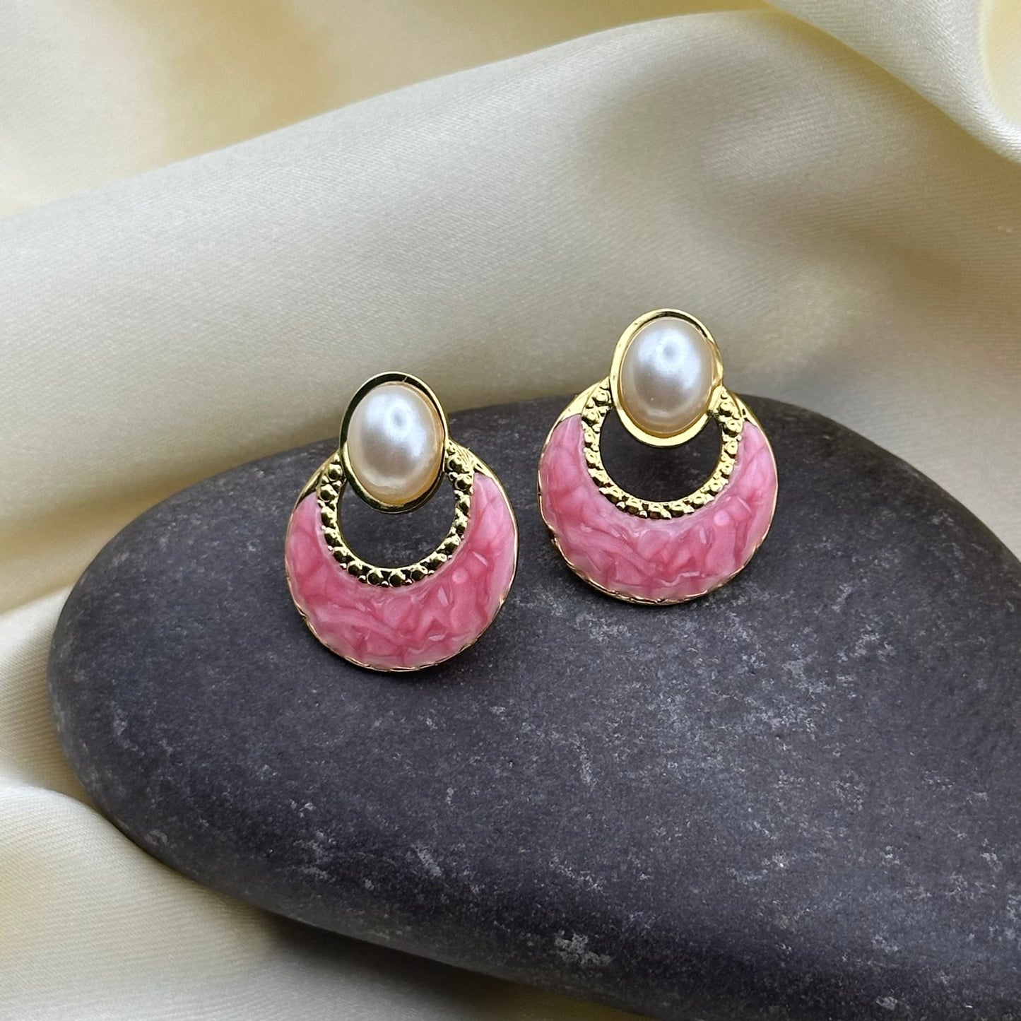 A pair of gold-tone hoop earrings with textured pink enamel and a white faux pearl at the top, displayed on a dark stone against a cream satin background.