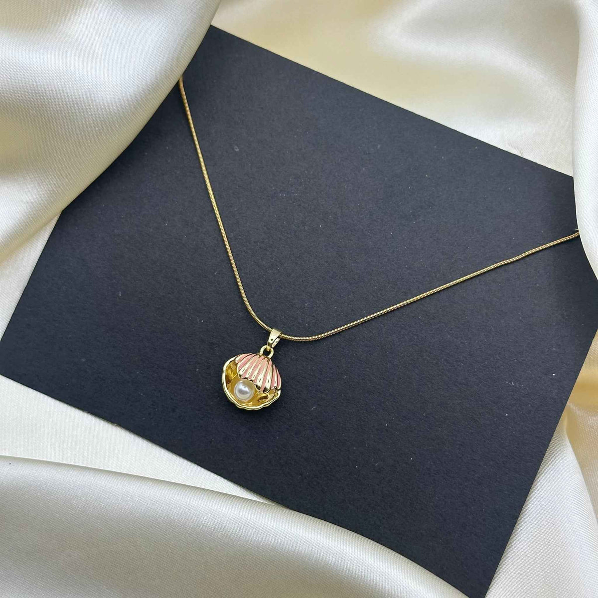 A pink enamel scallop shell pendant on a fine gold chain, displayed against a dark card background.