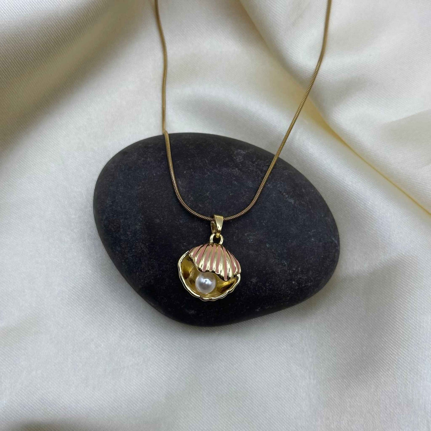 A gold seashell pendant necklace draped over a smooth grey pebble on cream satin.