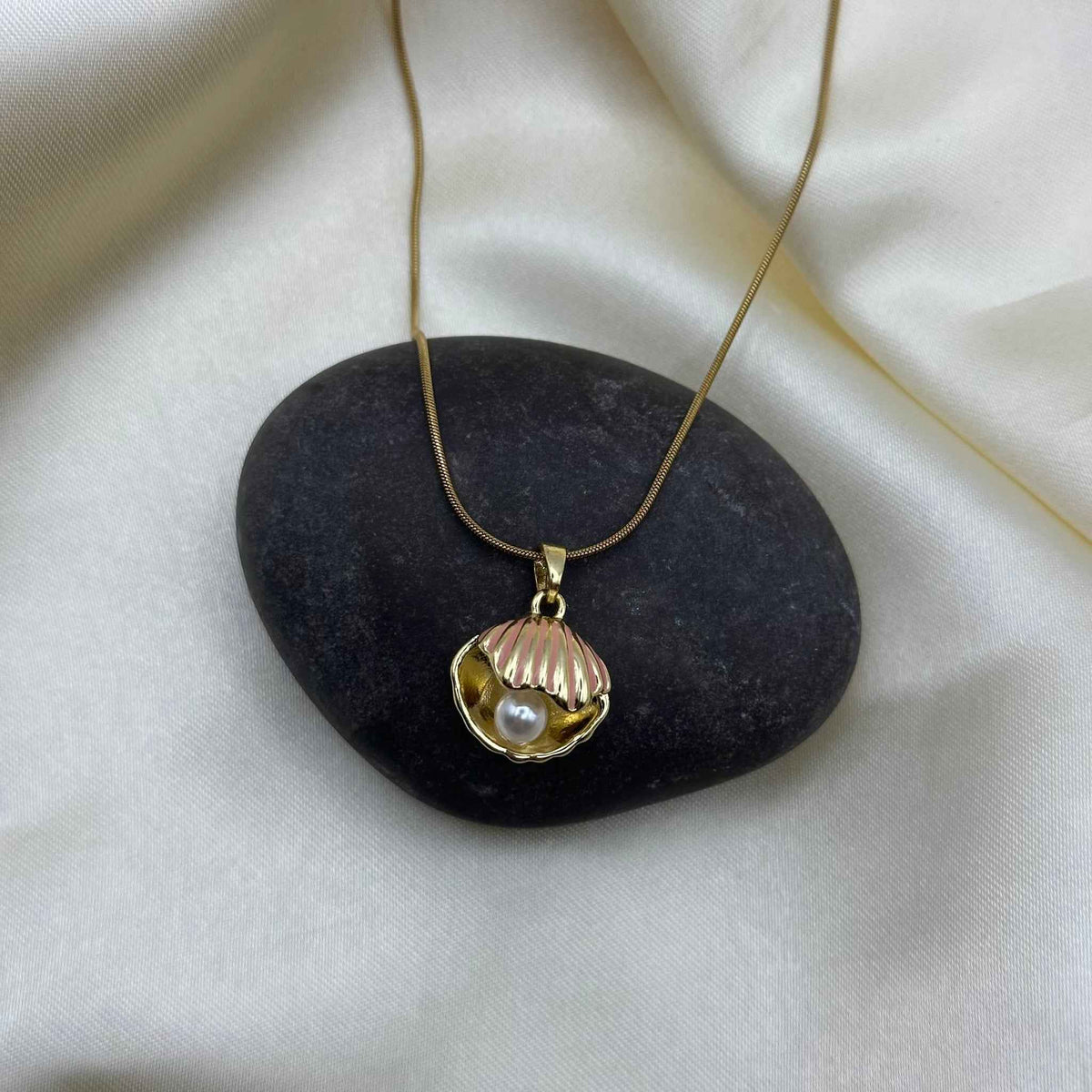 A gold seashell pendant necklace draped over a smooth grey pebble on cream satin.