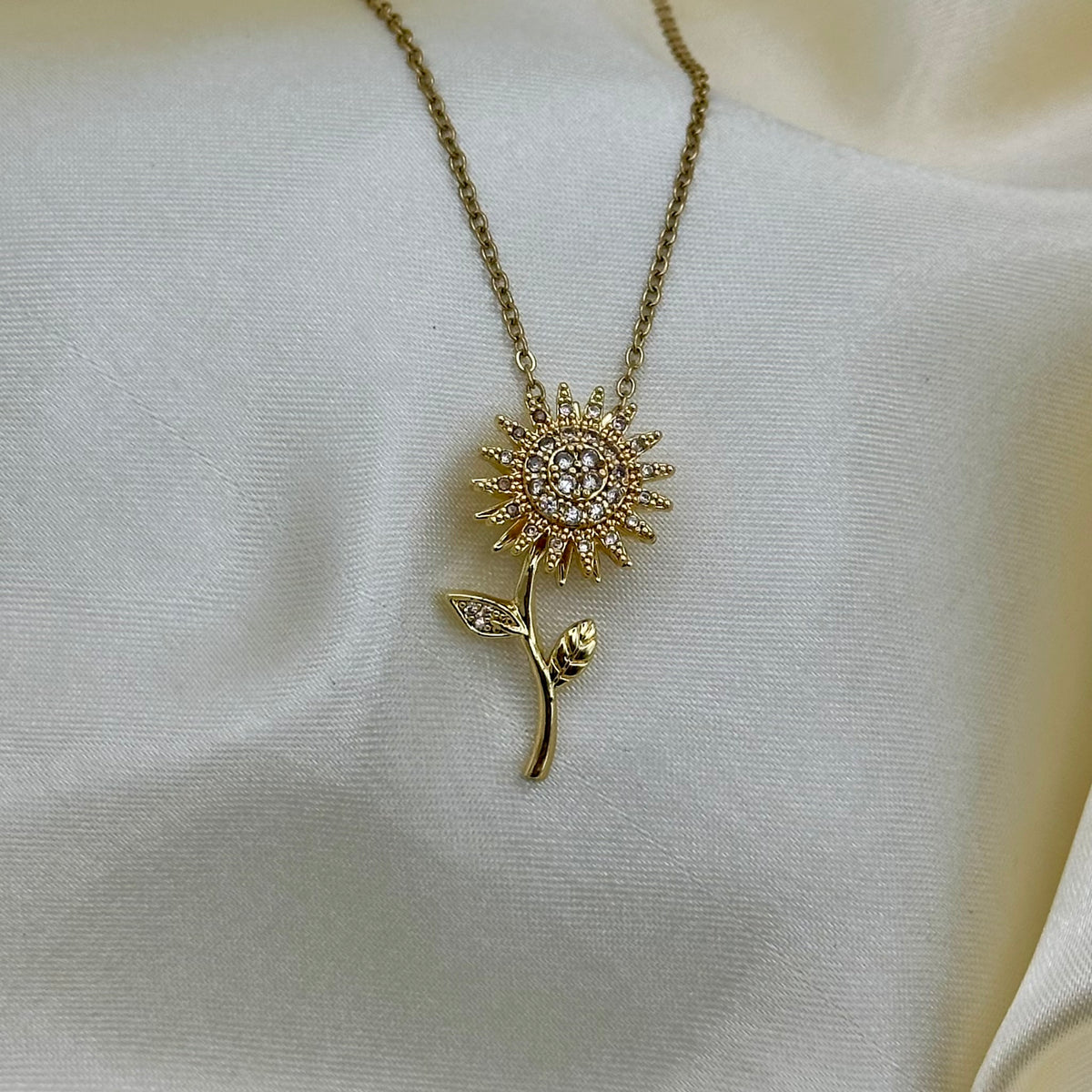 A gold-tone flower pendant necklace with clear rhinestones, displayed on draped cream satin fabric.