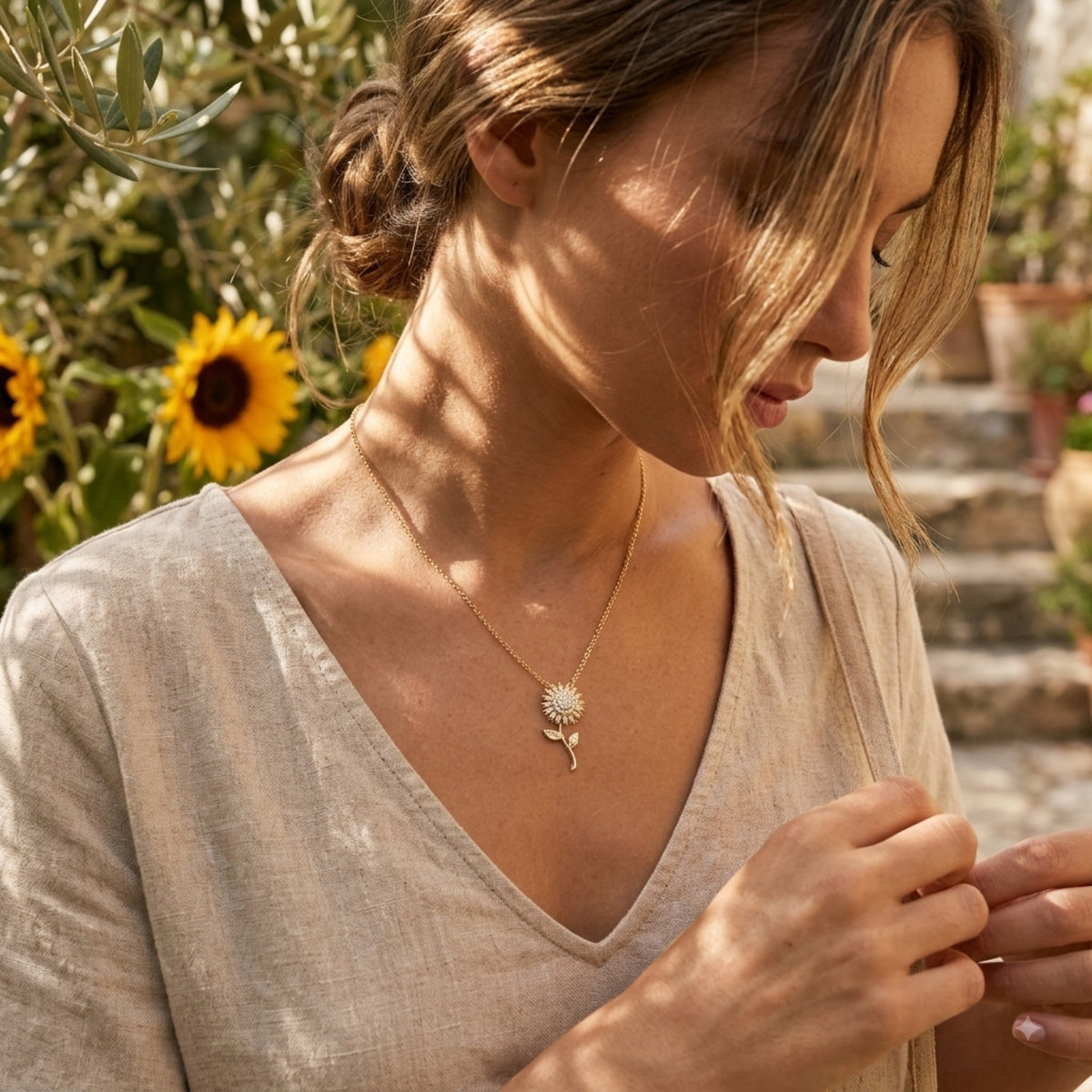 A model in a linen top wearing a gold sunflower pendant necklace, photographed outdoors with sunflowers in the background.