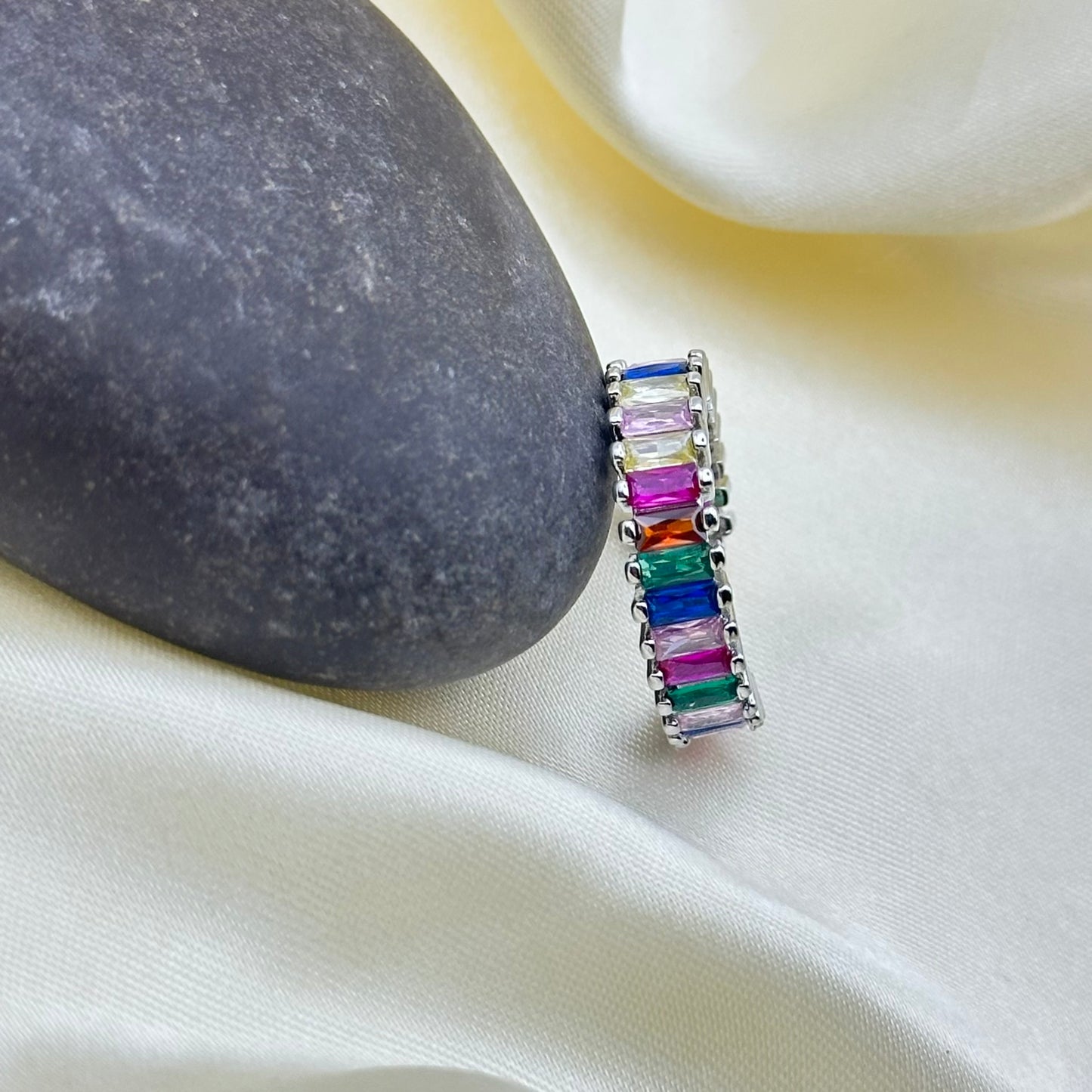 A silver ring featuring a vertical row of colorful rectangular gemstones in shades of pink, blue, green, orange, and purple, displayed on draped cream satin fabric next to a dark gray stone.