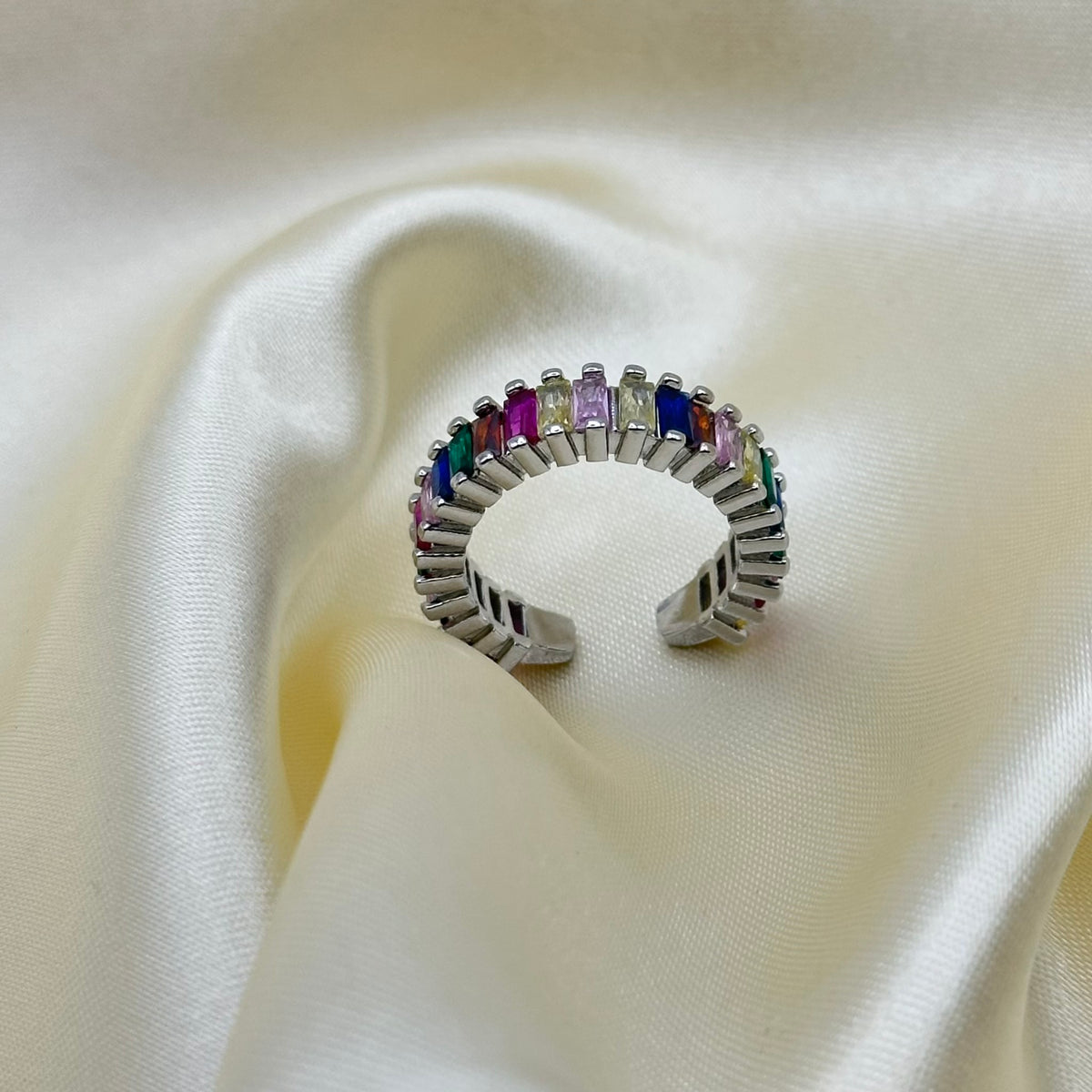 A silver-toned open cuff ring featuring a row of colorful rectangular gemstones in pink, purple, blue, green, and yellow, displayed on draped cream satin fabric.