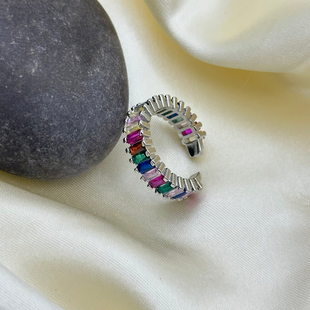 A silver-toned open cuff ring adorned with colorful rectangular gemstones in pink, orange, yellow, green, blue, and purple, styled on a cream satin fabric next to a dark gray stone.
