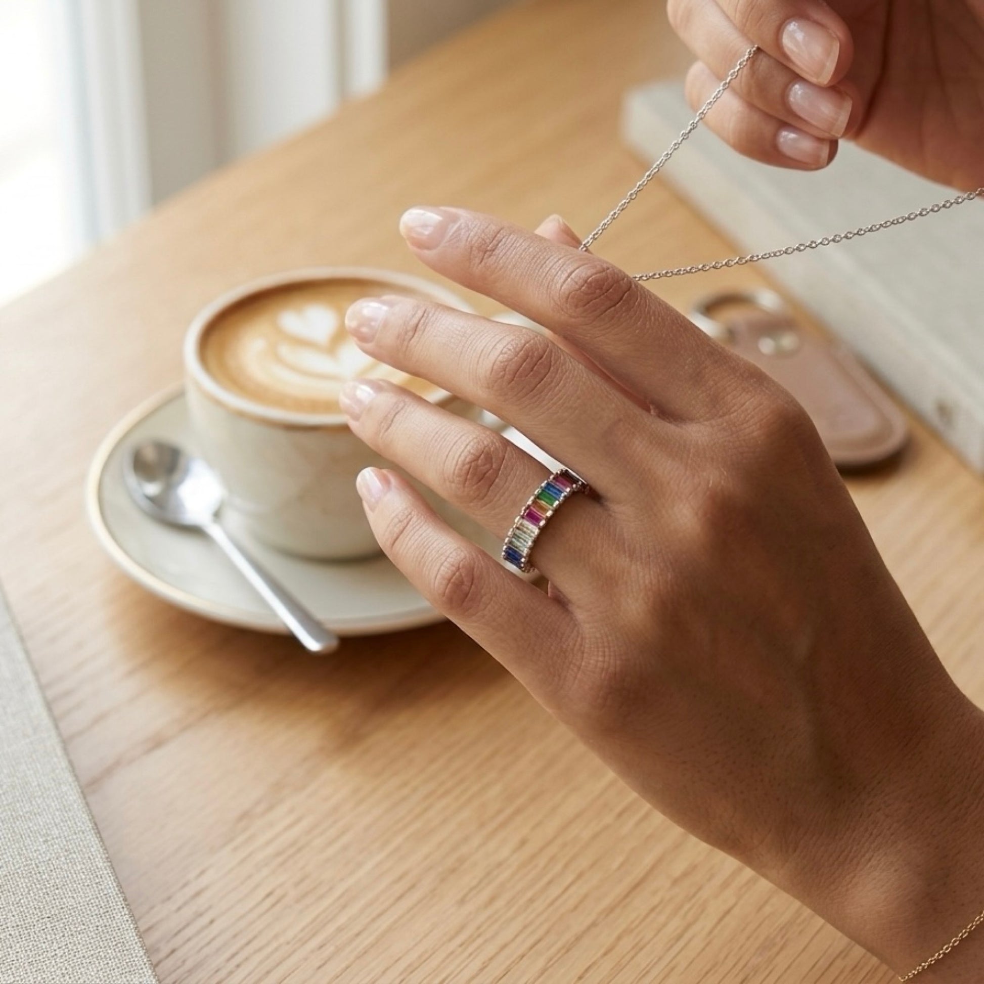 A close-up lifestyle shot of a person's hands holding a delicate silver chain necklace, with one hand wearing a silver band ring set with colorful gemstones. In the background, a cup of latte macchiato on a saucer sits on a light wooden table.