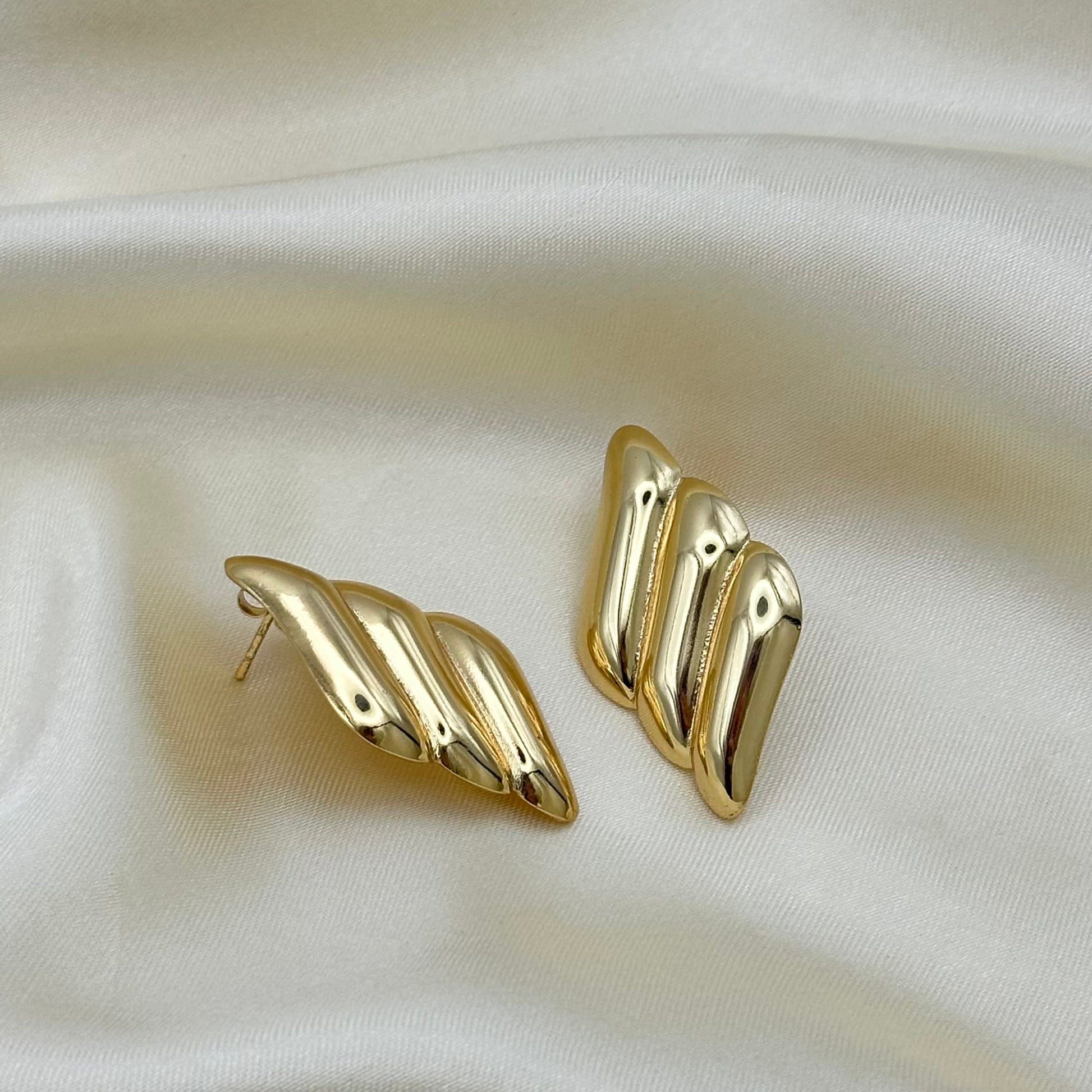 A pair of gold-toned, multi-lobed statement earrings displayed on a draped cream satin fabric background.