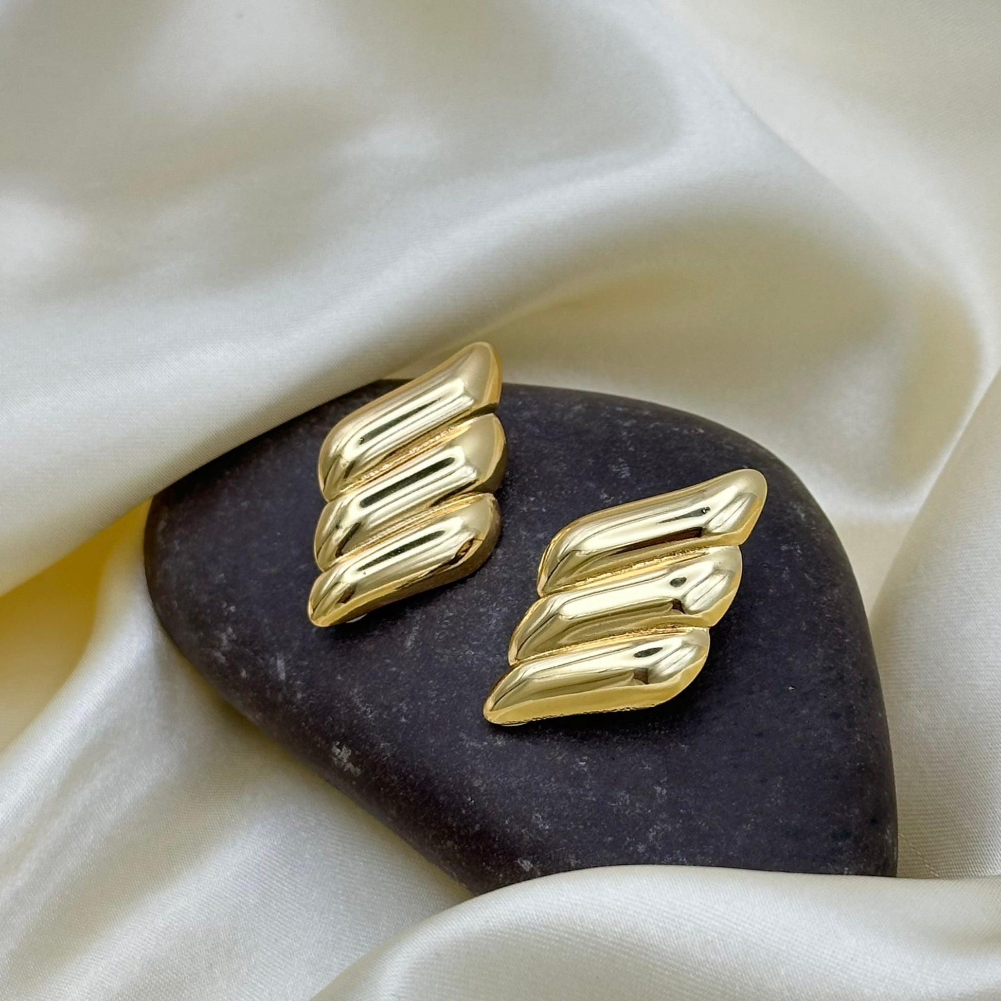 A pair of gold-toned, textured wing-shaped earrings displayed on a dark stone against a draped cream satin fabric background.