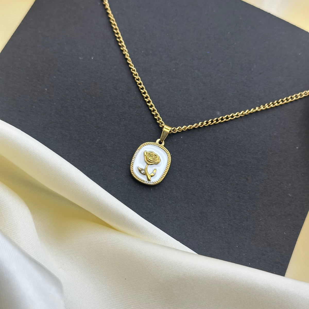 A gold chain necklace with a small oval pendant featuring a gold rose on a white enamel background, styled on a dark gray card with cream satin fabric.