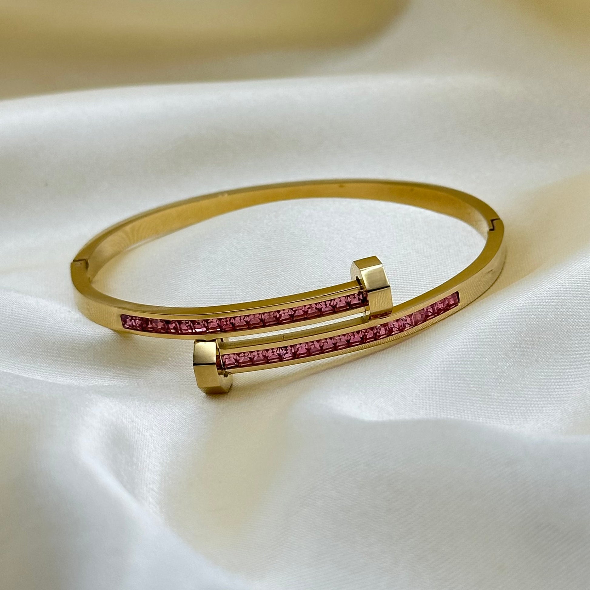 A gold bangle bracelet with two parallel bars, each accented by a channel of small pink gemstones, displayed on a draped white fabric background.