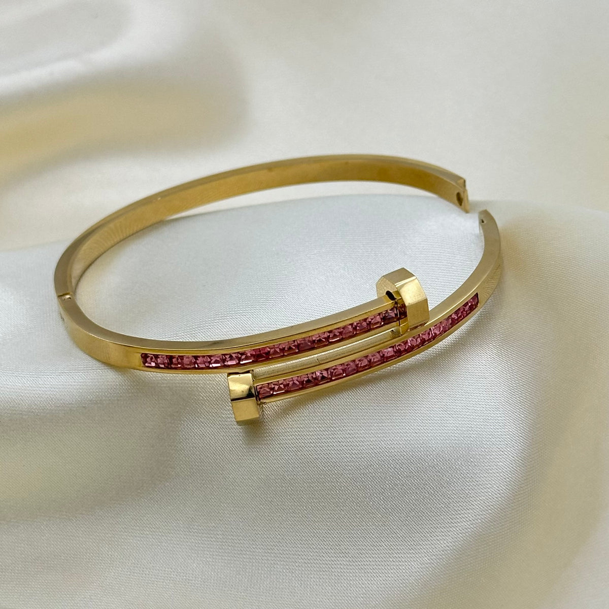 Gold-plated open cuff bangle bracelet with two parallel bars set with pink gemstones, displayed on a white satin fabric background.