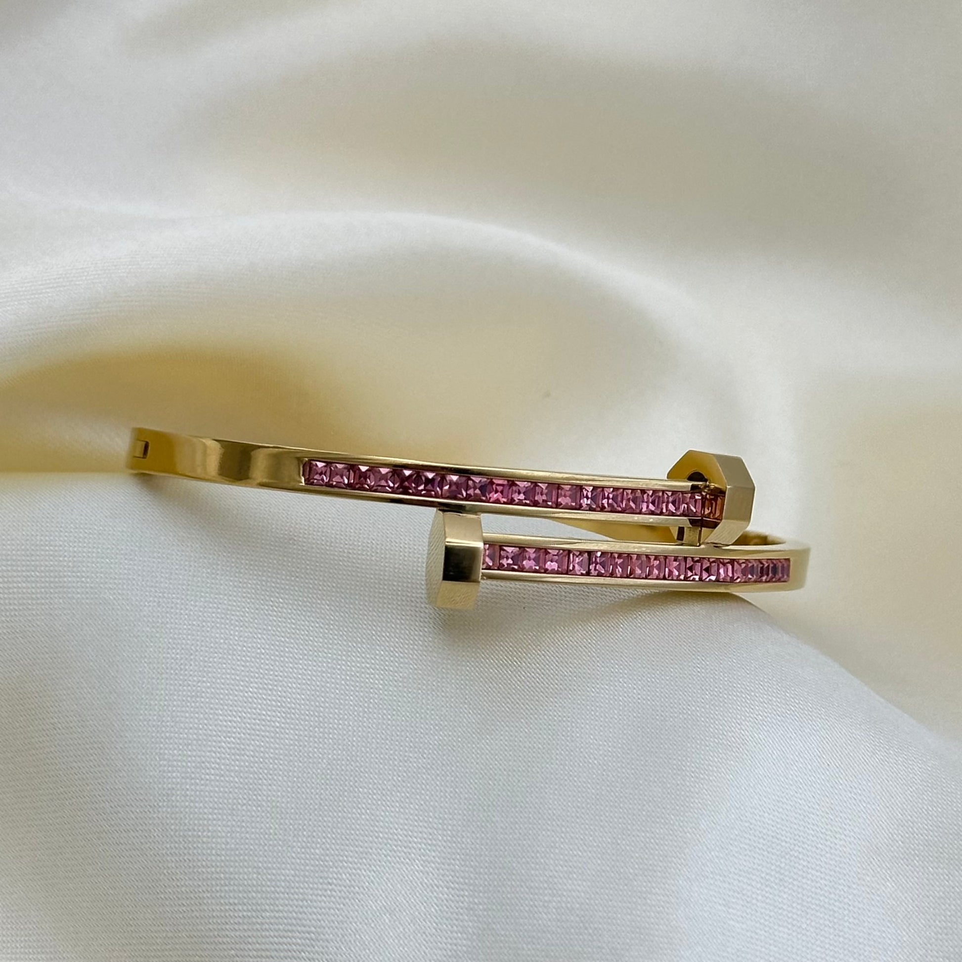 A gold-plated open cuff bangle bracelet with a row of pink gemstones, displayed on a draped white satin fabric.