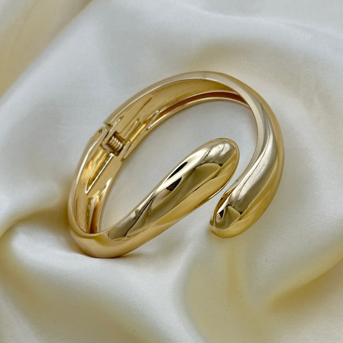 A polished gold-toned open cuff bracelet resting on draped cream satin fabric.