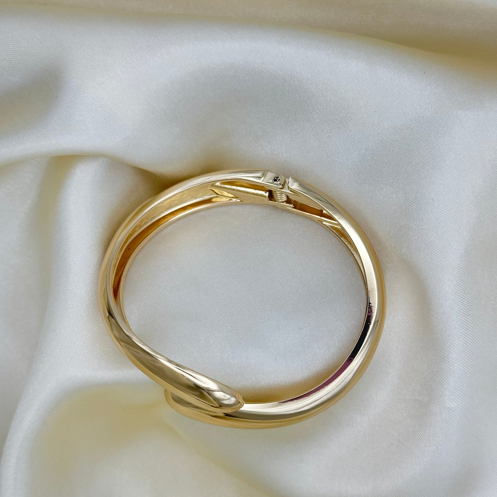 A polished gold bracelet with a twisted, open design resting on draped cream-colored satin fabric.