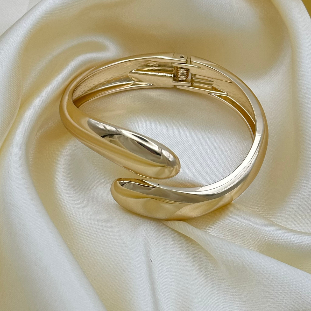 A polished gold-toned metal cuff bracelet with a sliding clasp, displayed on draped cream satin fabric.