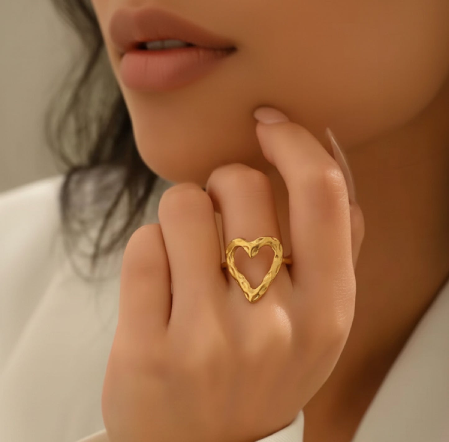A close-up lifestyle shot of a model wearing a textured, open-heart-shaped gold ring on her finger.