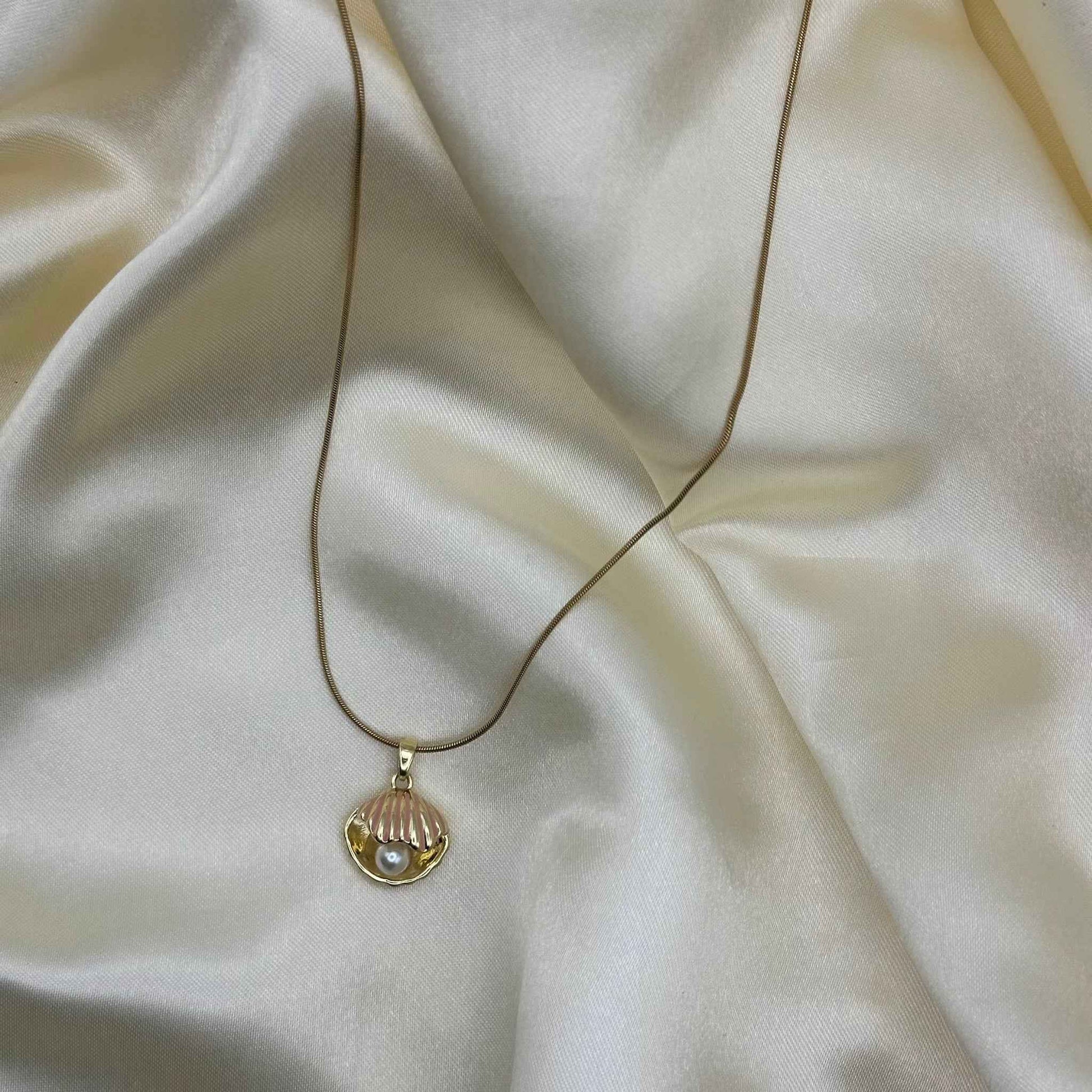 A gold seashell pendant necklace with pink enamel and a faux pearl, laid on soft cream satin.