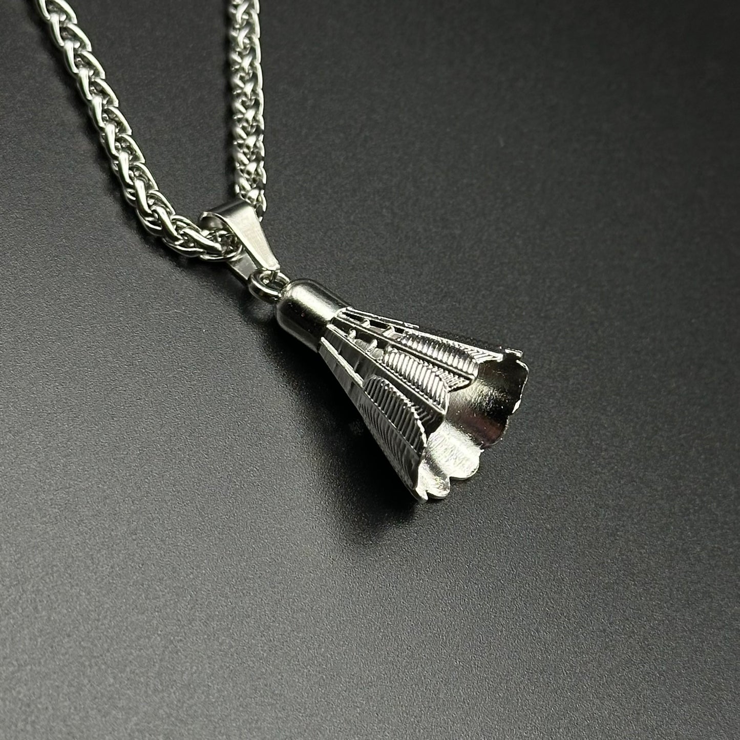 A silver-toned metal pendant necklace featuring a textured, shuttlecock design suspended from a matching silver chain against a dark gray background.