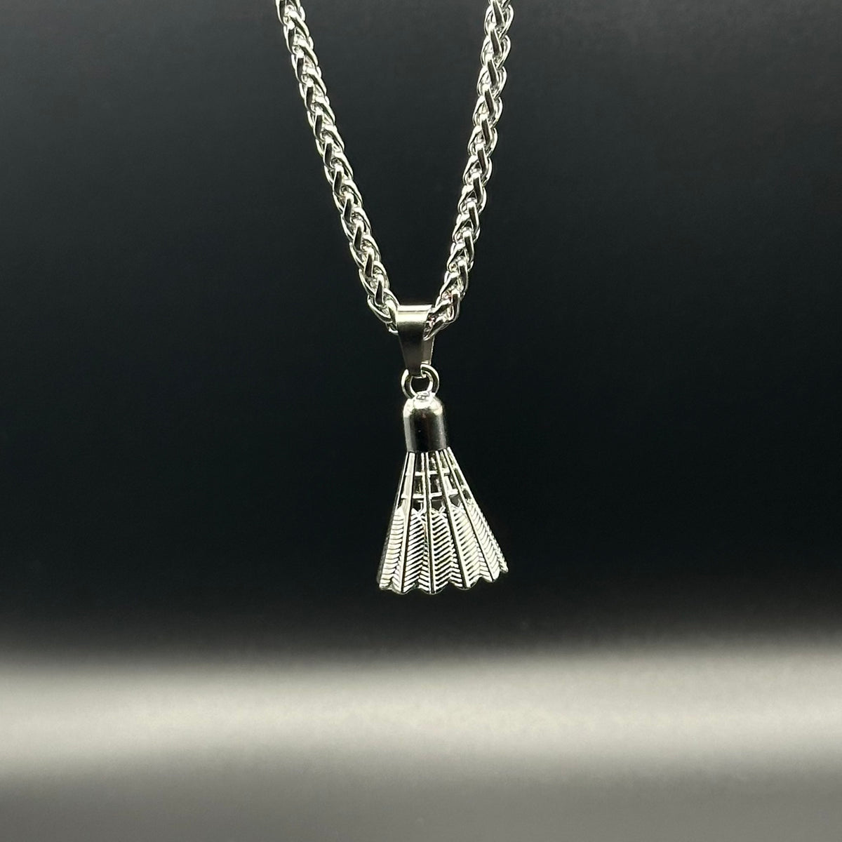 A silver chain necklace with a small shuttlecock-shaped pendant, shown against a dark gradient background.