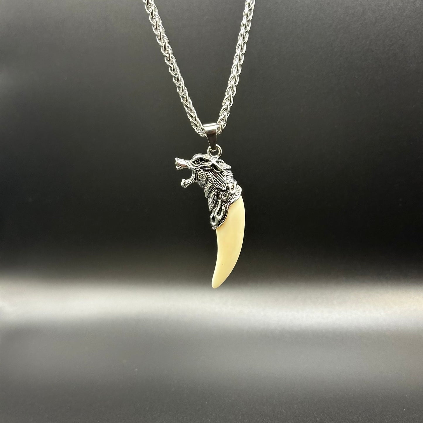 A silver wolf head pendant necklace with a curved ivory-colored tooth charm, displayed on a dark gray background.