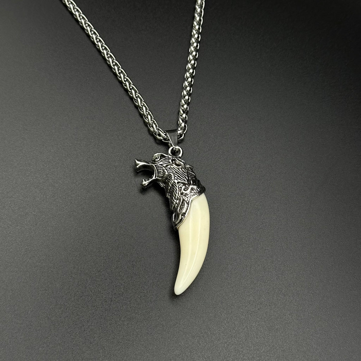 A wolf head pendant necklace with a cream-colored fang and a silver-toned head, hanging from a silver chain against a dark gray background.