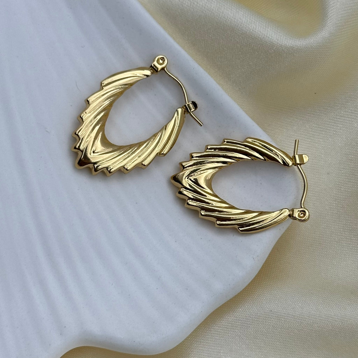 A pair of gold-toned textured hoop earrings with a layered, ribbed design, displayed on a white ceramic dish and cream-colored satin fabric.