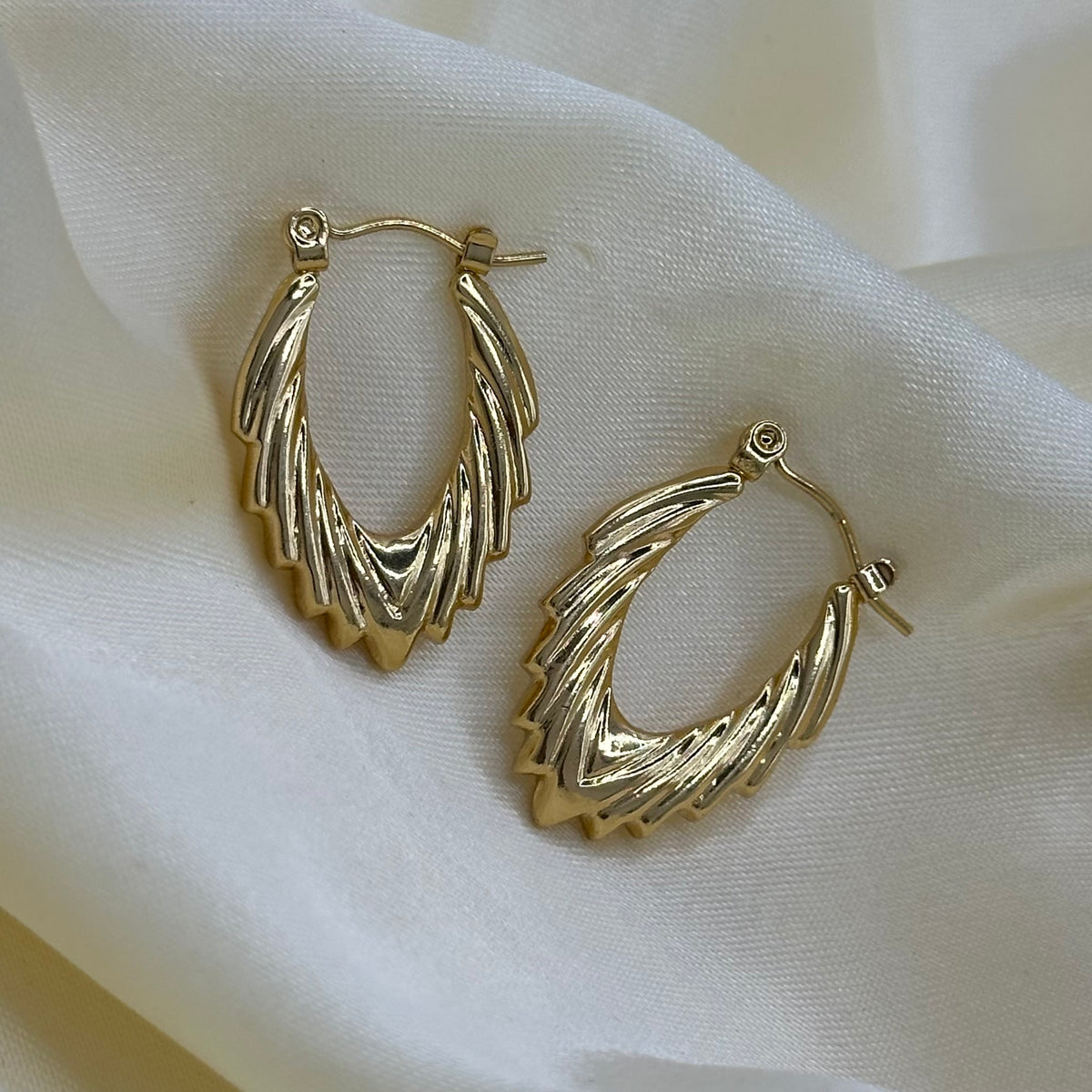 A pair of gold-toned hoop earrings with a textured, layered chevron design, displayed on a draped cream-colored fabric background.