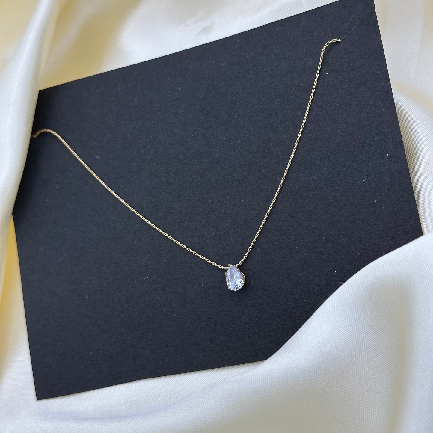 A delicate gold chain necklace with a small pear-cut clear gemstone pendant, displayed on a black card over draped white satin fabric.